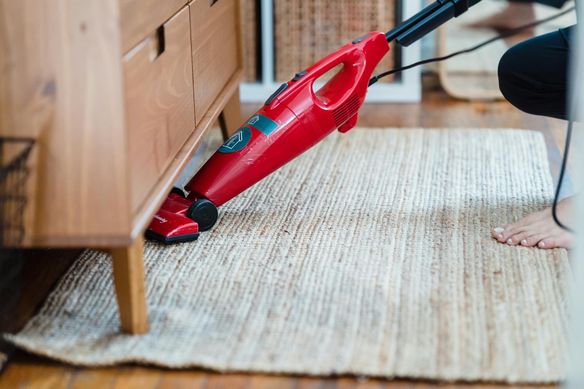 Person using a vacuum cleaner under furniture on a rug — thorough vacuuming is a key step in carpet stain prevention and preparation