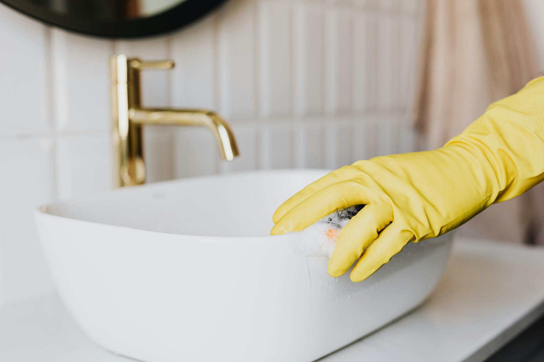Person in yellow rubber gloves cleaning a white bathroom sink