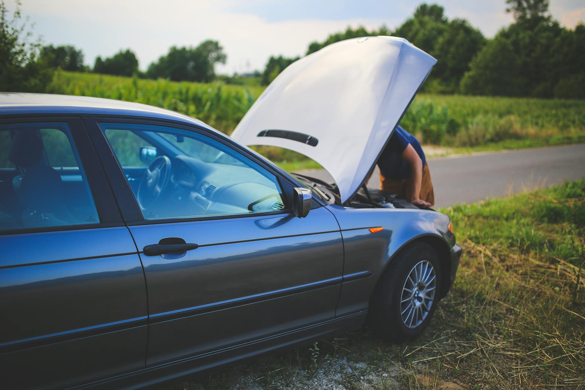 Man checking car engine with hood open by the side of a rural road