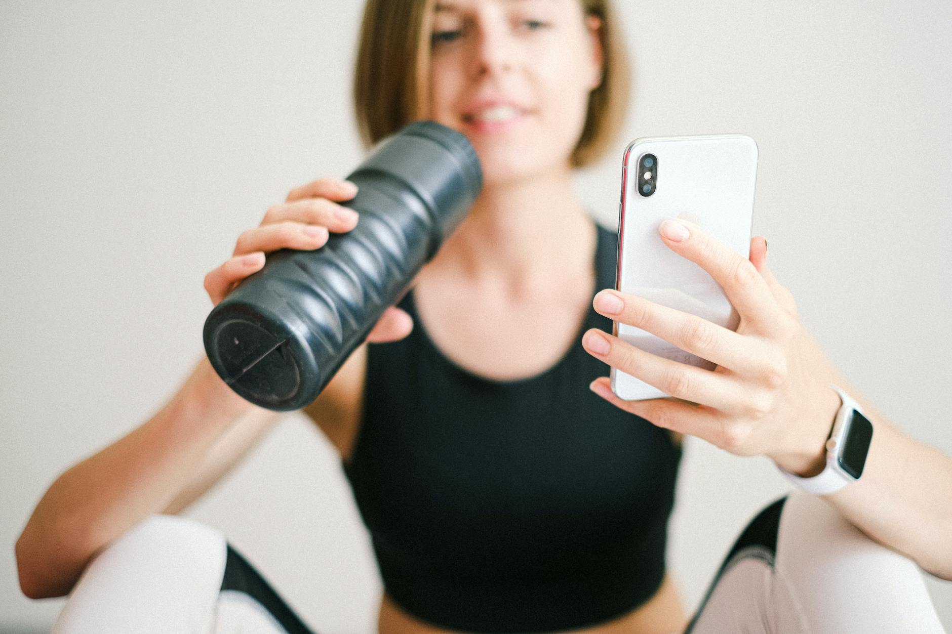 Woman checking smartphone while resting during a home workout with a sports bottle