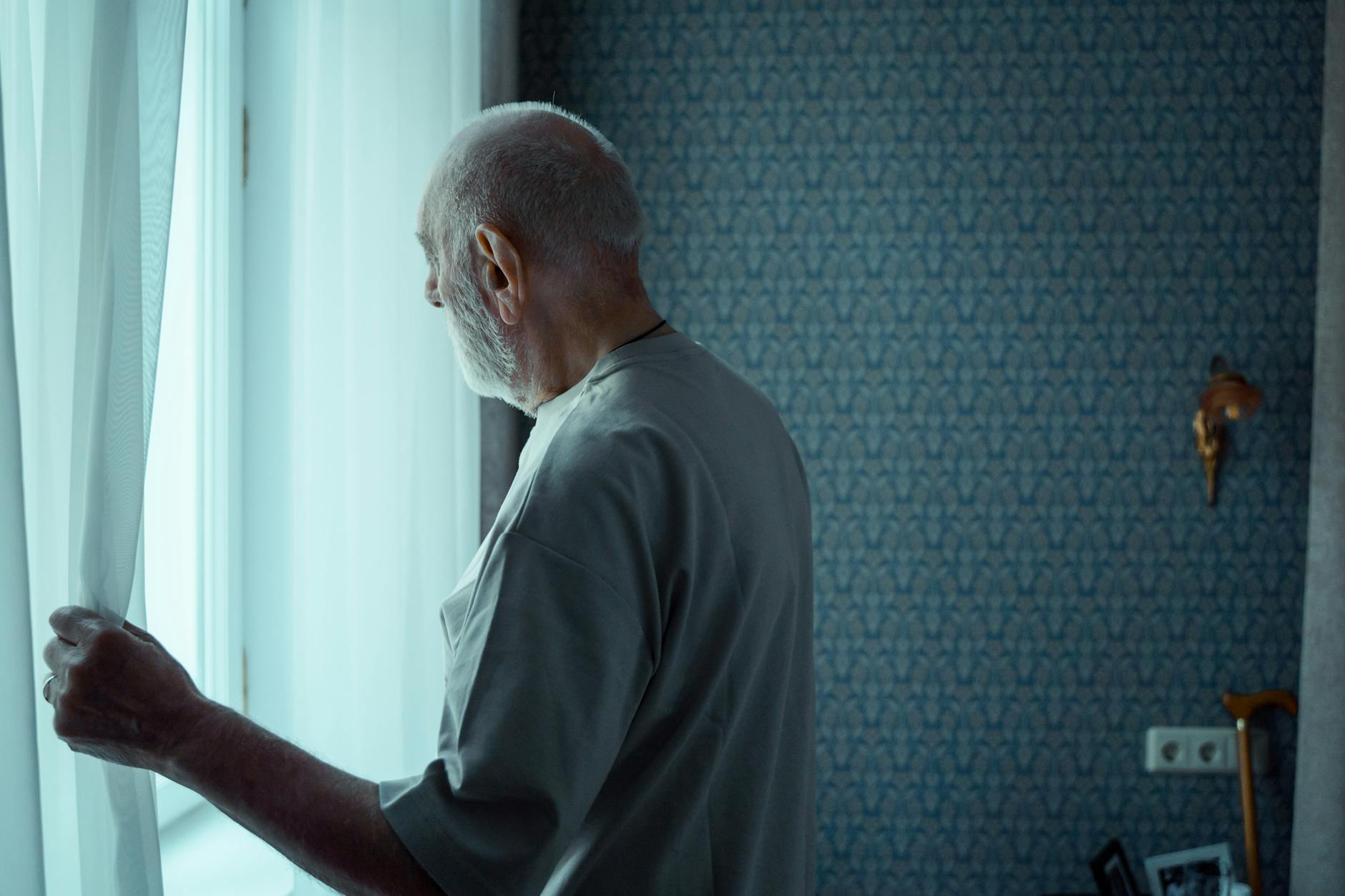 Senior man standing alone looking out a window, reflecting the social isolation that untreated hearing loss can cause